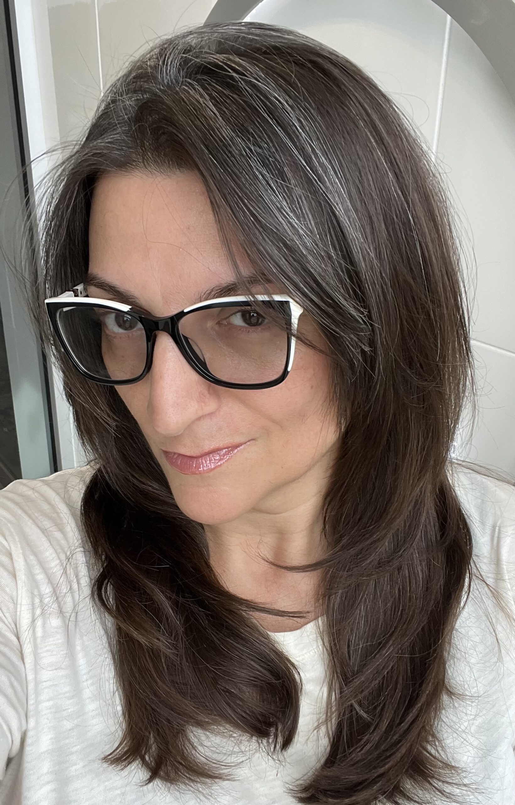 A light-skinned woman with long dark-brown hair with silver highlights, wearing a white top and fashionable black and white glasses. She has a slight smile and is looking directly into the camera.