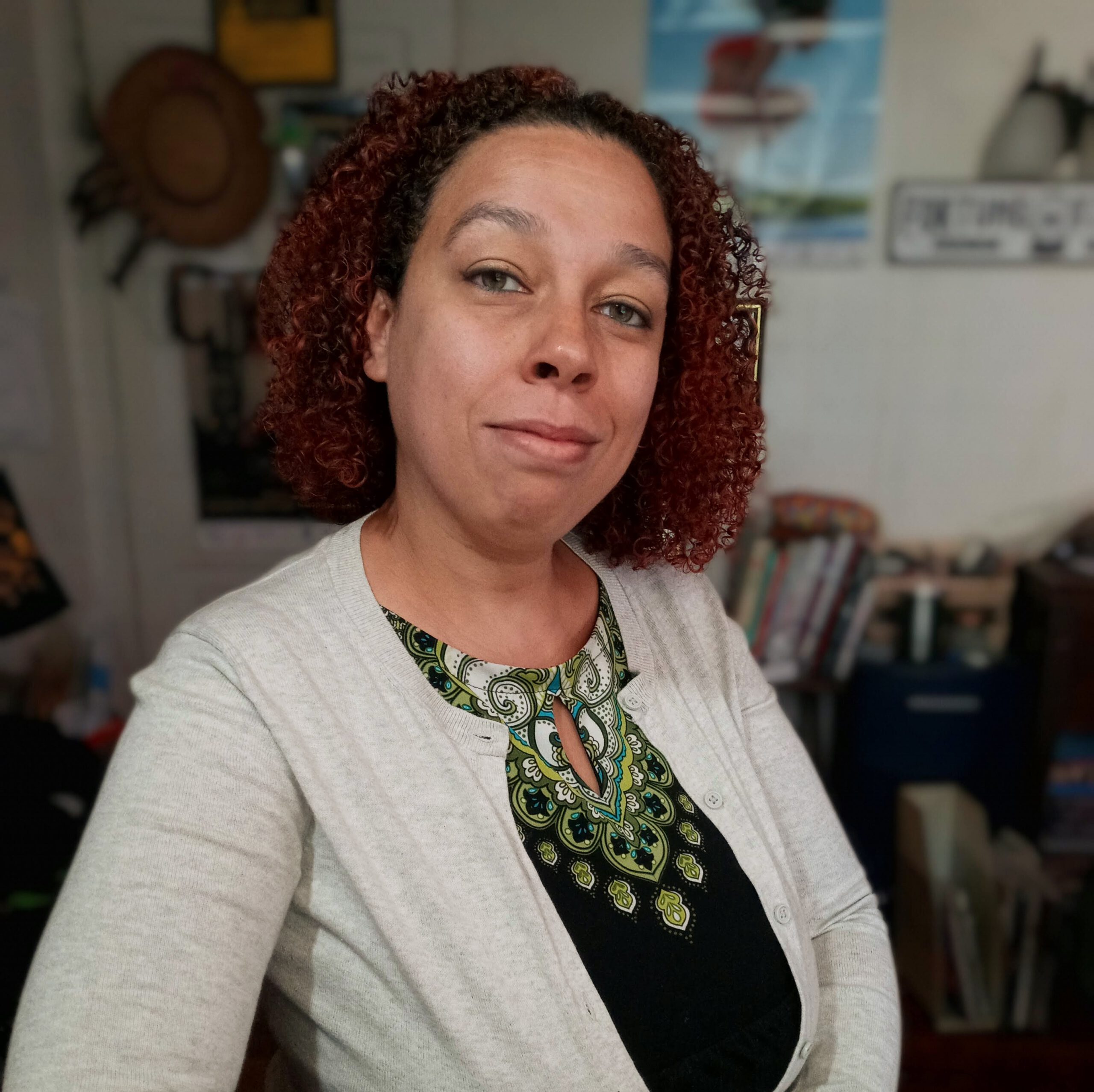 Tan skinned mixed race woman with brownish curly hair, wearing a black and green dress with a gray sweater in front of blurred background.