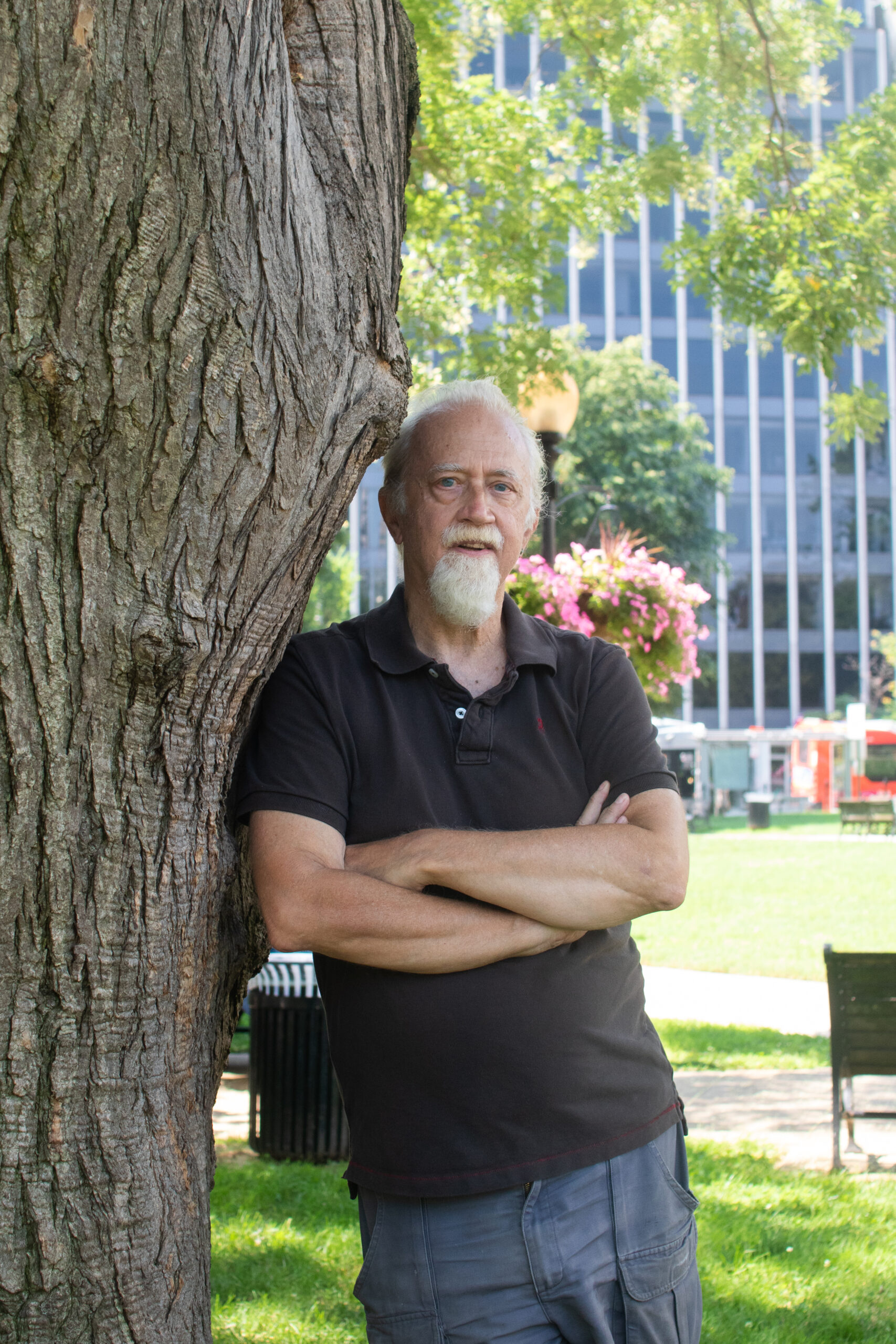 A white man with white hair and beard, wearing a black shirt, leans against a tree with arms
crossed.