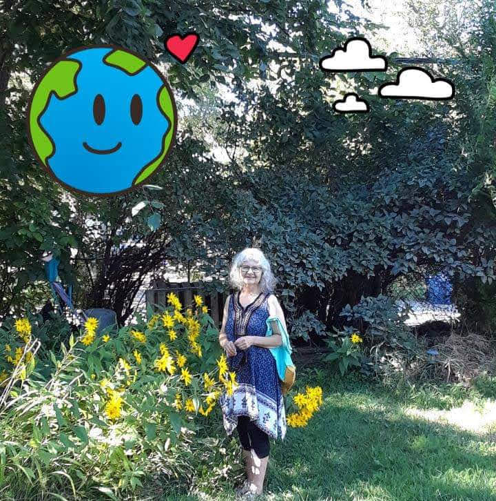 A light skinned grey haired smiling woman wearing a navy and white patterned top and black leggings is standing in her garden. Trees and bright yellow flowers tower over her.